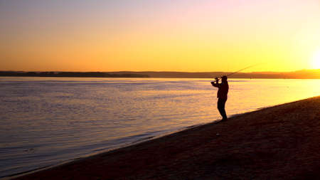 A man is fishing at sunrise by the river. The yellow sun rises from the horizon. Silhouette of a fisherman. A man throws a fishing rod.の写真素材