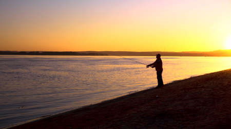 A man is fishing at sunrise by the river. The yellow sun rises from the horizon. Silhouette of a fisherman. A man throws a fishing rod.の写真素材