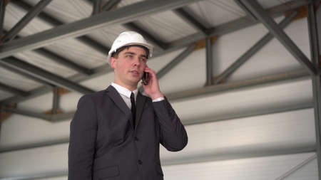 A young man in a helmet speaks on the phone at a construction site. The boss in a suit is talking on the phone.の写真素材