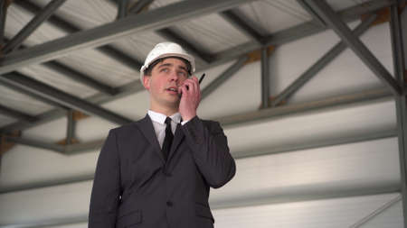 A young man in a helmet speaks on a walkie-talkie at a construction site. The boss in the suit looks around.の写真素材