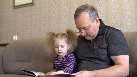 Granddaughter is reading a book with grandfather. The girl frowns at the book and listens carefully to Grandfather. Sitting on the couch together closeupの写真素材