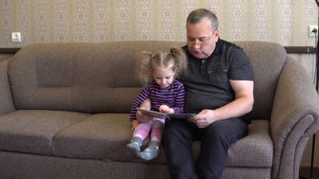 Granddaughter is reading a book with grandfather. The girl frowns at the book and listens carefully to Grandfather. Sitting on the couch together.の写真素材