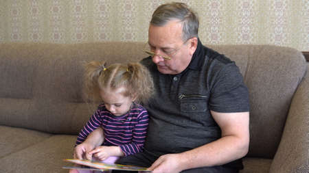 Granddaughter is reading a book with grandfather. The girl frowns at the book and listens carefully to Grandfather. Sitting on the couch together closeupの写真素材