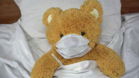 A teddy bear measures the temperature with a mercury thermometer. The bear lies in bed with a medical mask.の写真素材