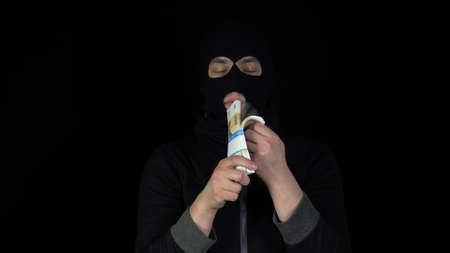 A man in a balaclava mask is standing with a bundle of rubles. The thug leafing through the money and rejoices. On a black background.の写真素材