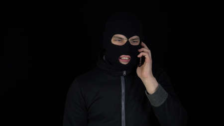 A man in a balaclava mask is talking on a phone. A thug screaming on the phone on a black background.の写真素材