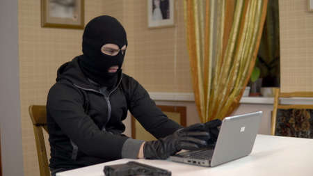 A robber is trying to hack into a laptop. A masked thug is sitting in a house and cannot hack into a laptop. Theft of data from a computer.の写真素材
