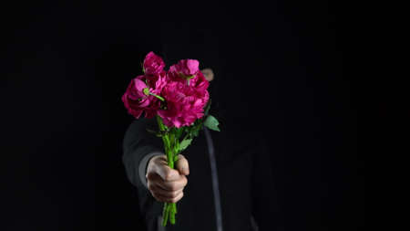 A man in a balaclava mask is standing with flowers. The bandit holds out a bouquet of pink flowers to the camera. On a black background.の写真素材