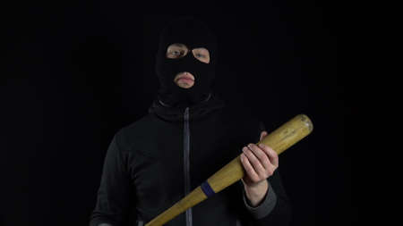 A man in a balaclava mask is standing with a baseball bat. A gangster is holding a baseball bat on a black background.の写真素材