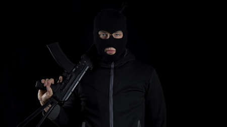A man in a balaclava mask stands with an AK-47 assault rifle. A bandit stands with a gun on his shoulder on a black background.の写真素材
