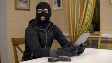 A robber is trying to hack into a tablet. A masked thug is sitting in a house and cannot hack into a tablet. Theft of data from a tablet.の写真素材