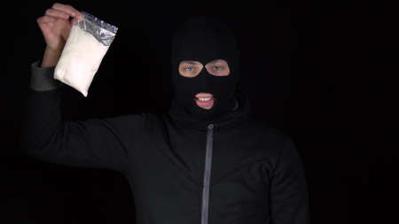 A man in a balaclava mask stands with a pack of heroin. The bandit holds a drug bag and rejoices. On a black background.の写真素材