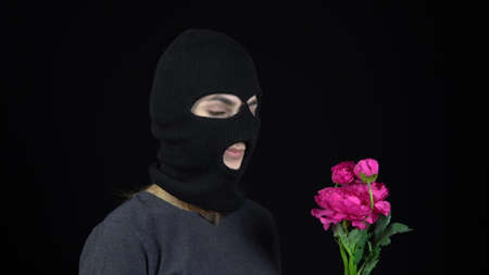 A Woman in a balaclava mask is standing with flowers. Bandit sniffs a bouquet of pink flowers on a black background.の写真素材