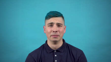 A young man in glasses with blue hair cries. Emotional man crying on a blue background.の写真素材