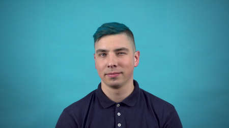 A young man in glasses with blue hair winks at his eyes. Emotional man winks on a blue background.の写真素材