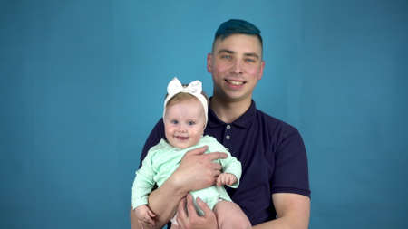 A young father with blue hair holds a baby in his arms. Alternative man with a child look at the...