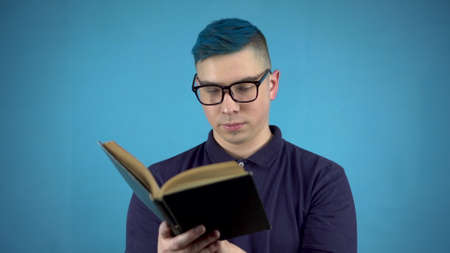 A young man in glasses with blue hair is reading a book. Alternative man with a book in hands on a...