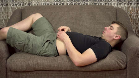 A young man lies on a sofa touching his fat belly. Gained excess weight in quarantine.の写真素材