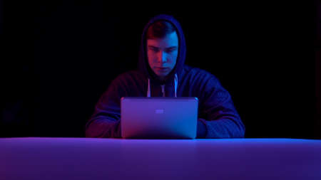 Young man in a hood with a laptop. Hacker makes a hack through a laptop. Blue and red light falls on...