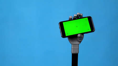 A mechanical hand holds a phone with a green screen. Gray cyborg hand holding a smartphone on a blue background. Chroma key.の写真素材