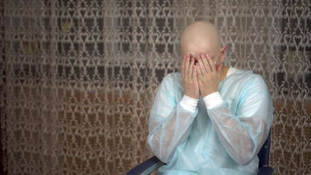 A bald young man with cancer looks at the camera and cries. The patient covers his face with his...