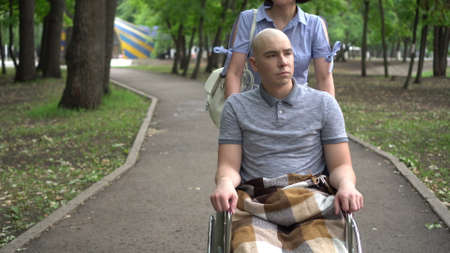 A woman is driving a young man with oncology in a wheelchair through the park. The man is bald due...