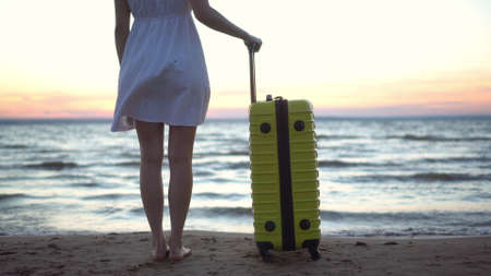 Young woman with a yellow suitcase on the beach by the sea. A girl in a white dress looks at the sunset close-up.の写真素材