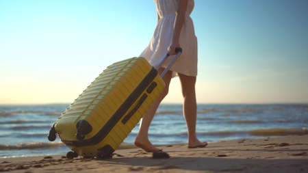Young woman walks along the beach of the sea with a yellow suitcase closeup. A girl in a white dress walks barefoot on the sand.の写真素材