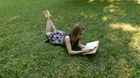 A young girl in a dress lies on the green grass and reads a book.の写真素材