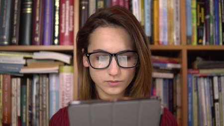 A young woman is reading a book in a tablet. A woman with glasses carefully looks at the tablet. In the background are books on bookshelves. Book libraryの写真素材