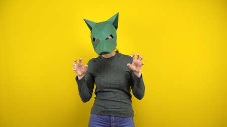 A young woman in a cardboard jackal mask depicts an animal and looks at the camera on a yellow...