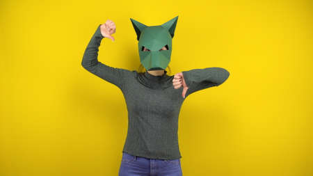 Young woman in a cardboard jackal mask shows dislikes with thumbs on a yellow background. Woman in...