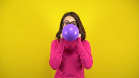A young woman inflates a purple balloon with her mouth on a yellow background. Girl in a pink turtleneck and glasses.の写真素材