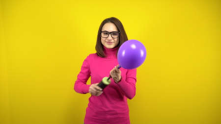 A young woman inflates a purple balloon with a pump on a yellow background. Girl in a pink turtleneck and glasses.の写真素材