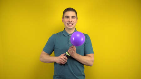 A young man inflates a purple balloon with a pump on a yellow background. Man in a green polo.の写真素材