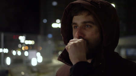 A young man stands in the cold in the evening outside and warms his hands with his breath.の写真素材