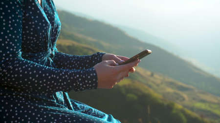 A young woman in a dress sits with a phone in her hands against a background of mountains closeup. The girl travels in the Caucasus mountains.の写真素材