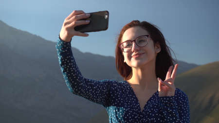 A young woman in a dress stands in the mountains and takes a selfie on a smartphone.の写真素材