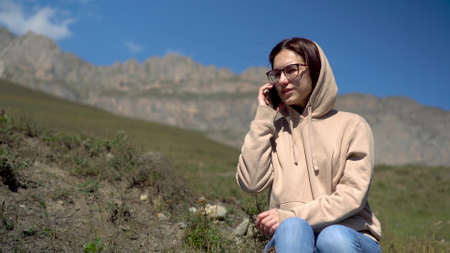 A young woman in a sweatshirt sits in the mountains and speaks on the phone. The girl travels in the...