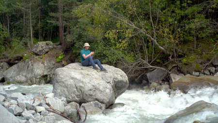 A young man in a Panama hat sits on a stone with a phone in his hands in the mountains by the river.の写真素材