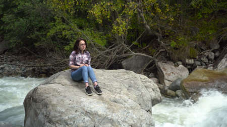 A young woman with glasses sits on a stone with headphones in the mountains near the river. Travel to the mountains.の写真素材