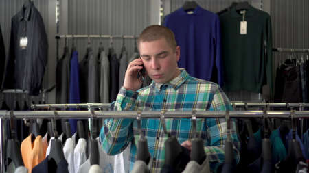 A young man chooses clothes in a store and speaks on the phone. Clothing store.