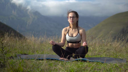 Young woman in tracksuit practices yoga performing lotus position in the mountains. The camera moves creating a parallax effect.の写真素材