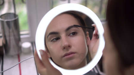 A young woman looks into a small illuminated mirror and paints her eyelashes with mascara. View of the girl through the mirror.の写真素材