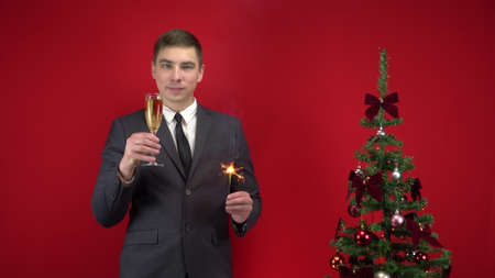 A young man stands with a glass of champagne and a sparkler near the Christmas tree on a red background. A man in a formal suit.の写真素材