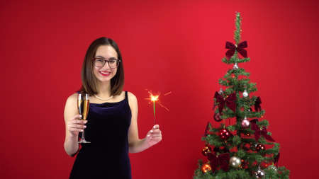 A young woman stands with a glass of champagne and a sparkler near the Christmas tree on a red background. A girl with glasses and an evening dress.の写真素材