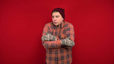 A young man in a red shirt, hat and mittens is cold and hugs himself to keep warm. Shooting in the...
