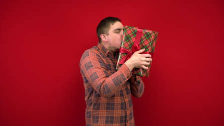 A young man shakes a Christmas present in his hands and rejoices. Shooting in the studio on a red background.の写真素材