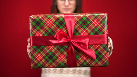 A young woman holds out a Christmas gift in the camera. Shooting in the studio on a red background.の写真素材