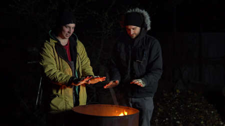 Two homeless young men bask in the fire. Men stand near a barrel of fire at night.の写真素材